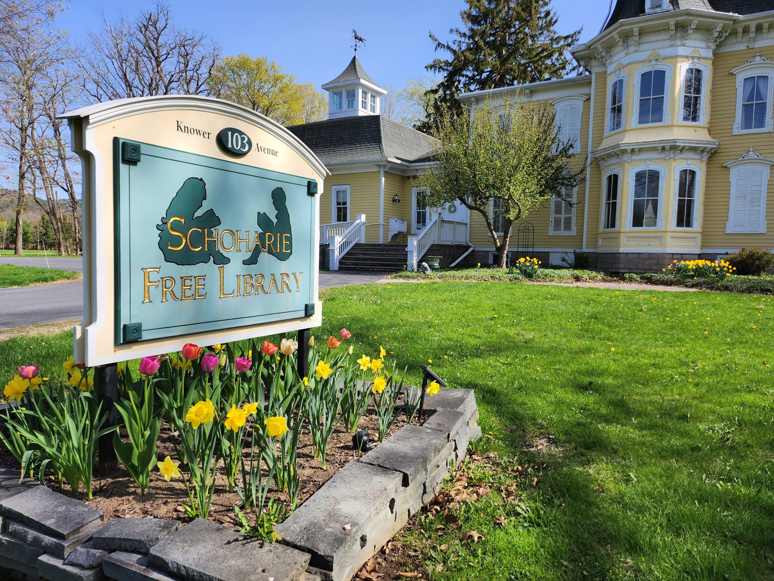 SHO - Library Bridge street view Schoharie Free Library sign in a tulip bed with the Schoharie Free Library in the background.